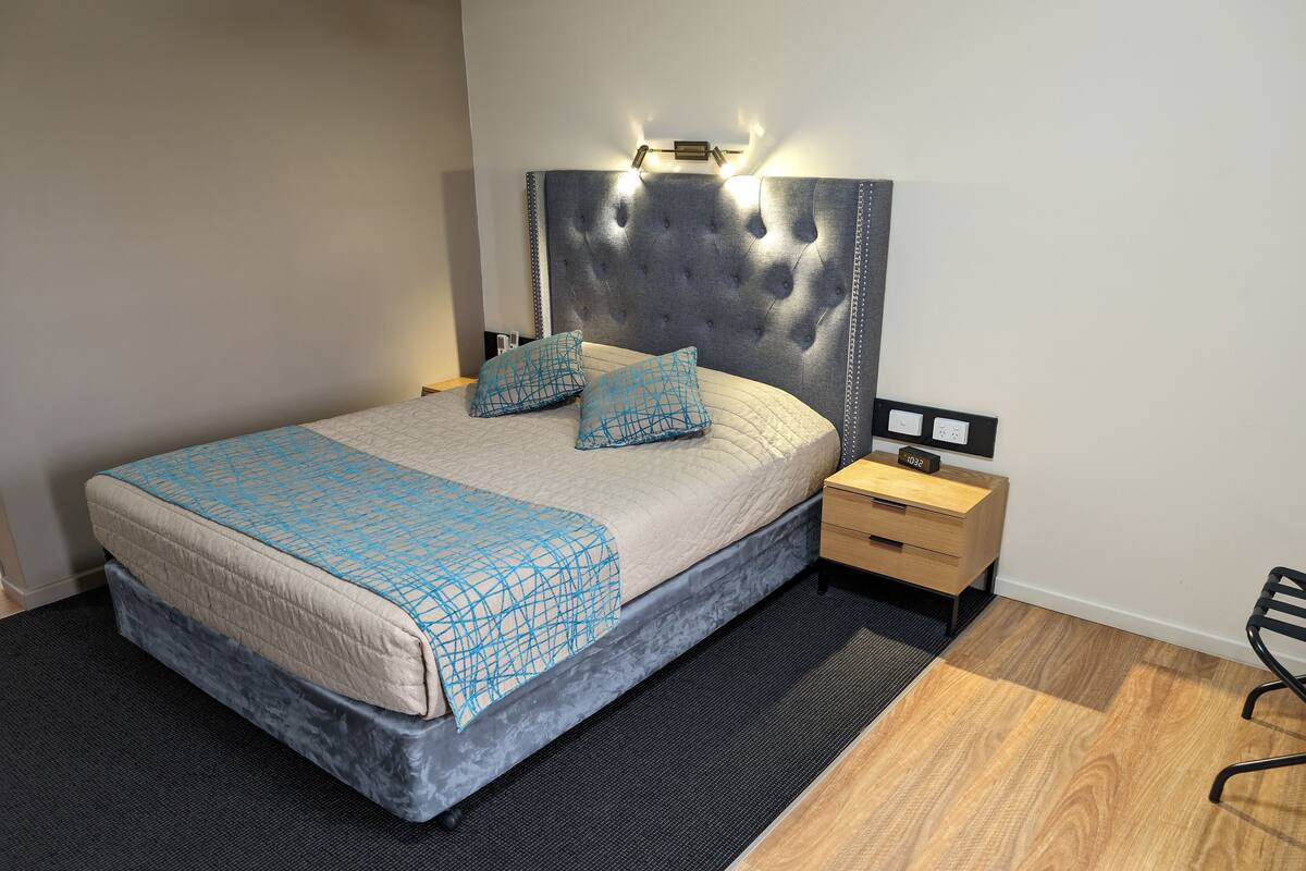 A modern bedroom with a tufted headboard, a bed with a quilted cover, two matching pillows, a bedside table with a clock, and a chair.