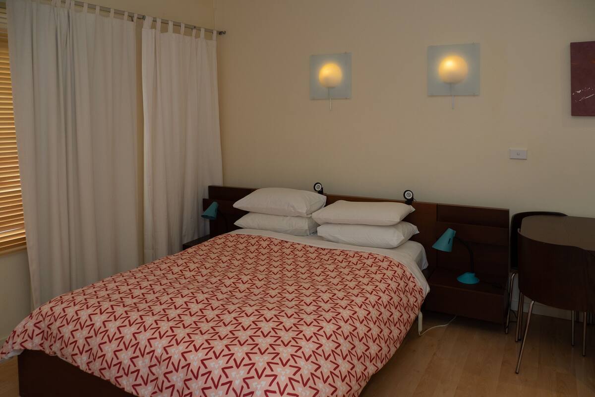 A neatly made bed with a patterned bedspread, a wooden headboard, two bedside lamps, and a small table with a chair.