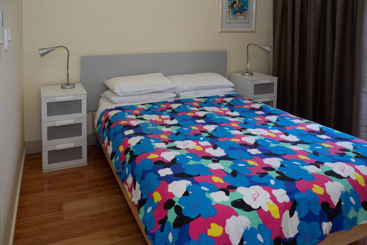 A bedroom with a bed, two bedside tables, a nightstand, a wall-mounted lamp, and a framed picture.