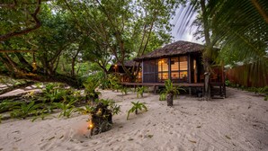 Una estructura con techo de paja y terraza de madera, rodeada de exuberante follaje tropical y una playa de arena.