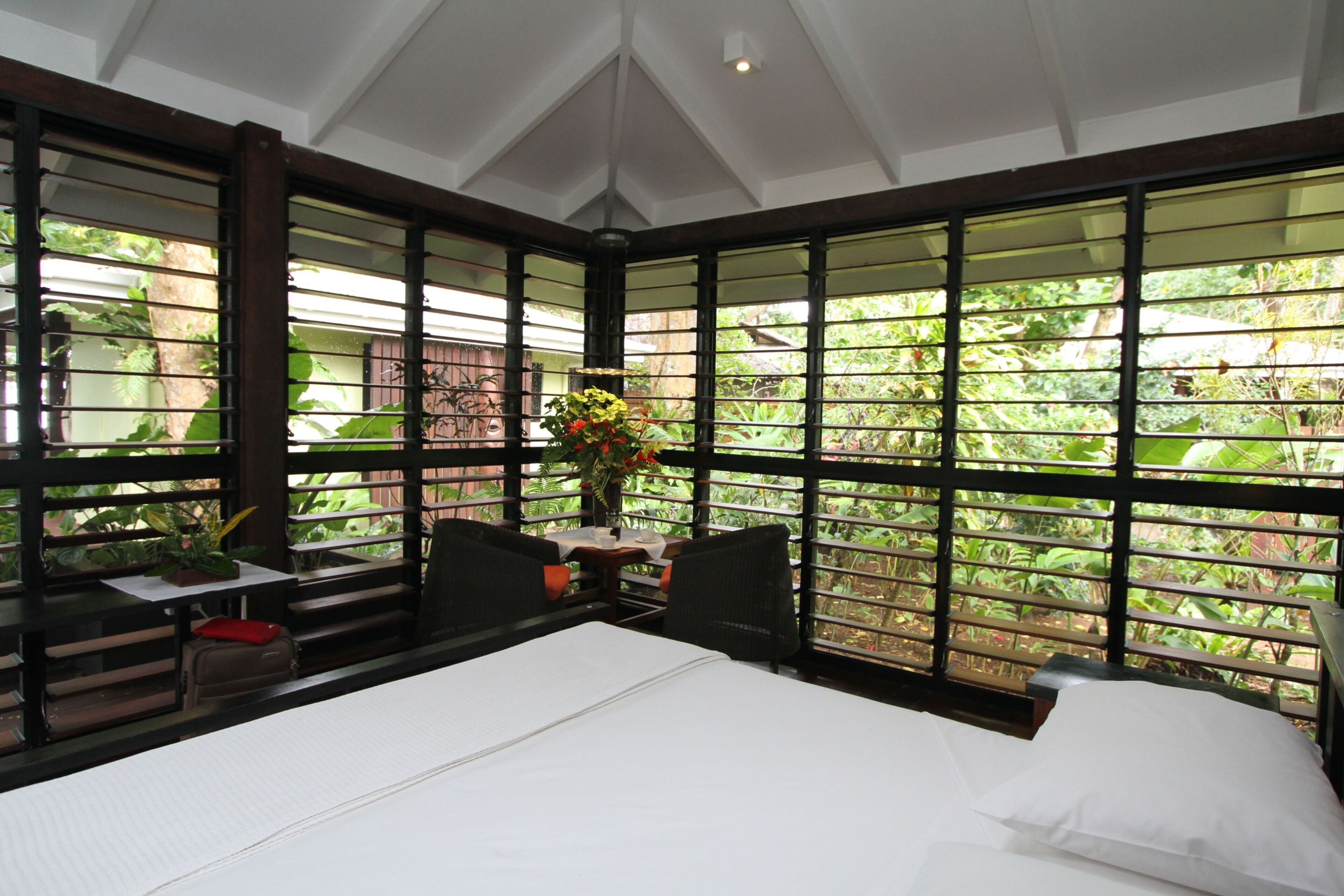 A room with a bed, a table with chairs, and a window with a view of greenery.