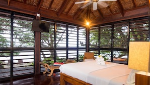 Una habitación con una cama, un banco de madera, un altavoz y grandes ventanales con vistas a una playa.