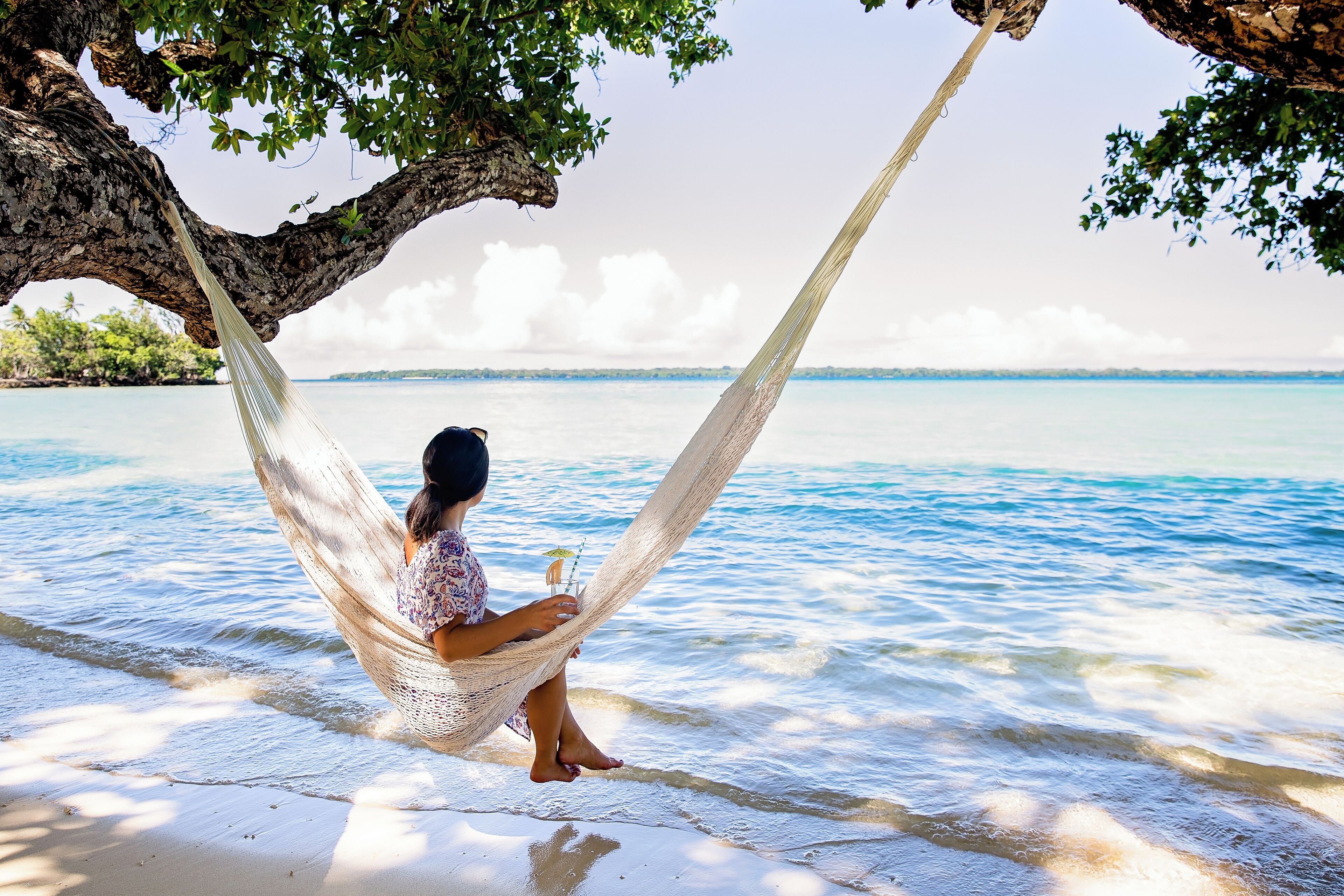 A hammock tied between two trees on a beach with clear blue water in the background.