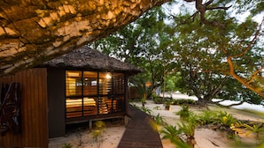Una estructura de madera con techo de paja, rodeada de arena y plantas tropicales, comunicada por una pasarela de madera.