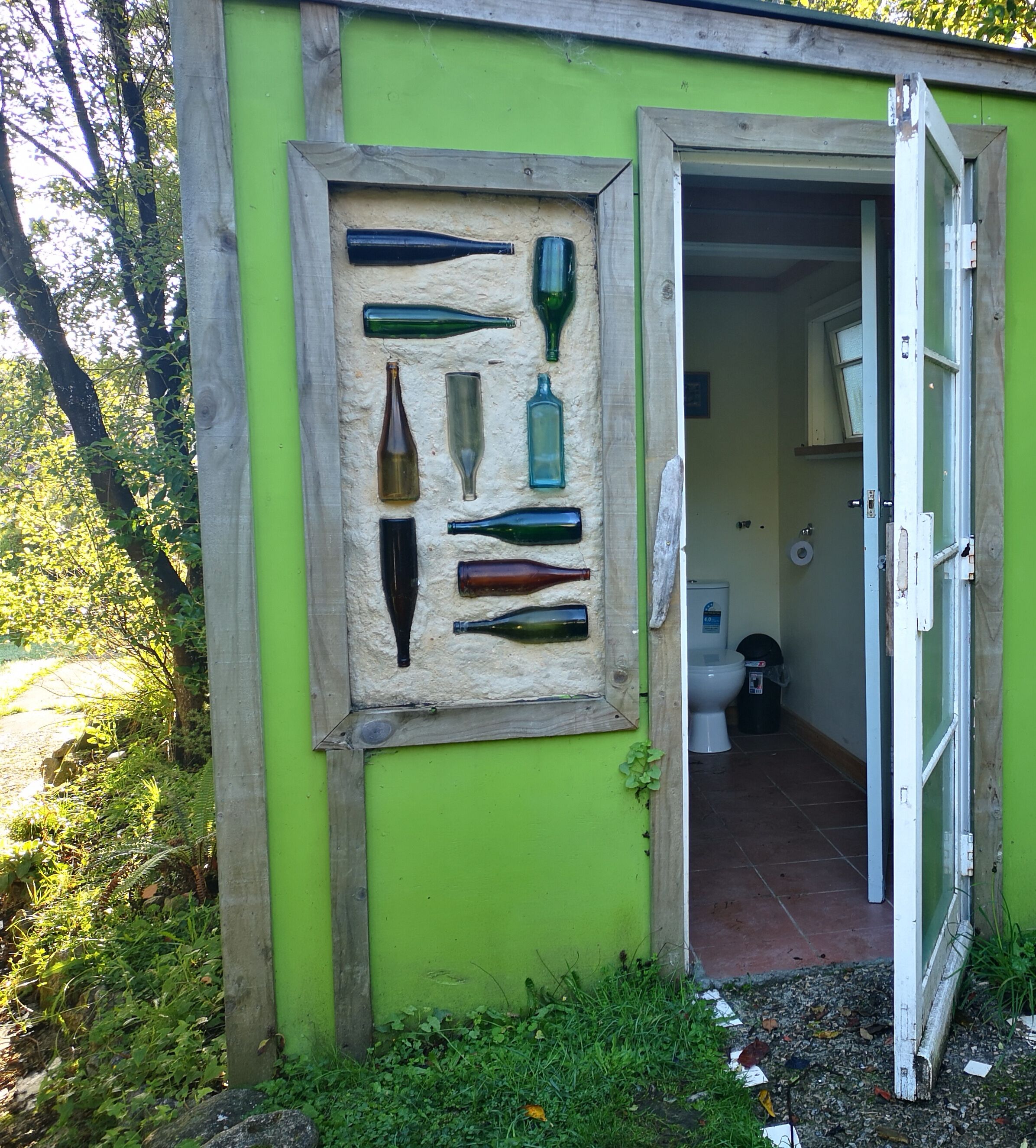Un edificio pequeño y verde con una puerta que da a un baño. Se observa una vitrina empotrada en la pared con varias botellas de vidrio.