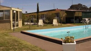 Piscina con sillas de descanso y una zona cubierta, rodeada por un césped y un edificio con techo rojo.