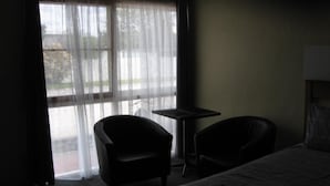 Habitación de hotel con cama, dos sillas, una mesita y una ventana con cortinas translúcidas.