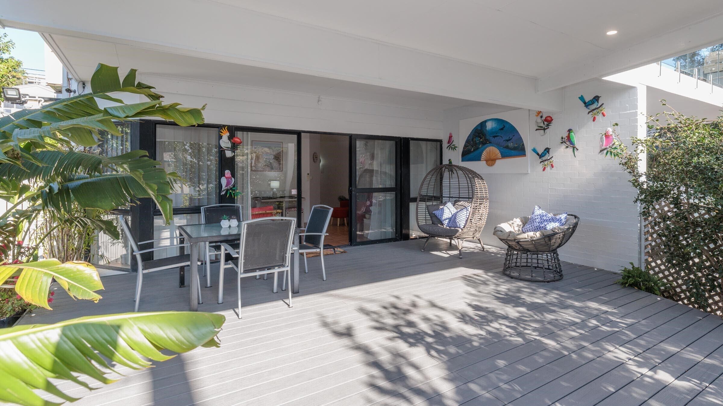 A patio with a dining table and chairs, a wicker chair, and a decorative wall with bird and flower art.