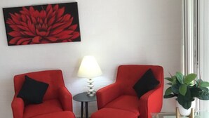 Un salon avec deux fauteuils rouges, une petite table ronde surmontée d’une lampe, et un grand tableau floral rouge accroché au mur.