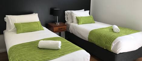 Ein Hotelzimmer mit zwei Betten, jeweils mit weißer Bettwäsche und grünen Akzenten. Dazwischen befindet sich ein Nachttisch mit einer Lampe.