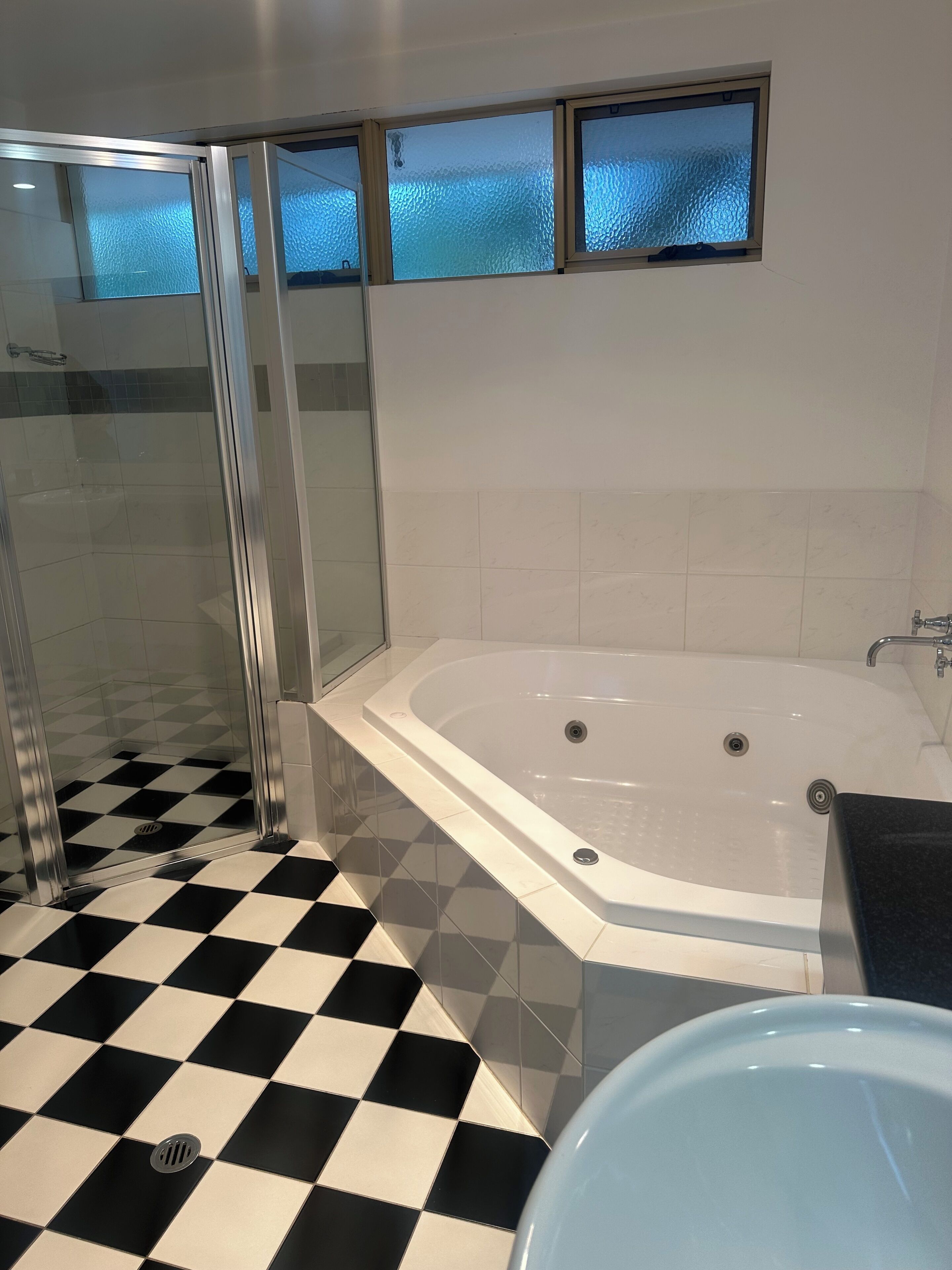 Un baño con piso a cuadros, ducha con cerramiento de vidrio, bañera blanca y ventana con vidrio esmerilado.