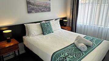 Habitación de hotel con cama, mesita de noche, lámpara, un cuadro en la pared y ventana con cortinas.