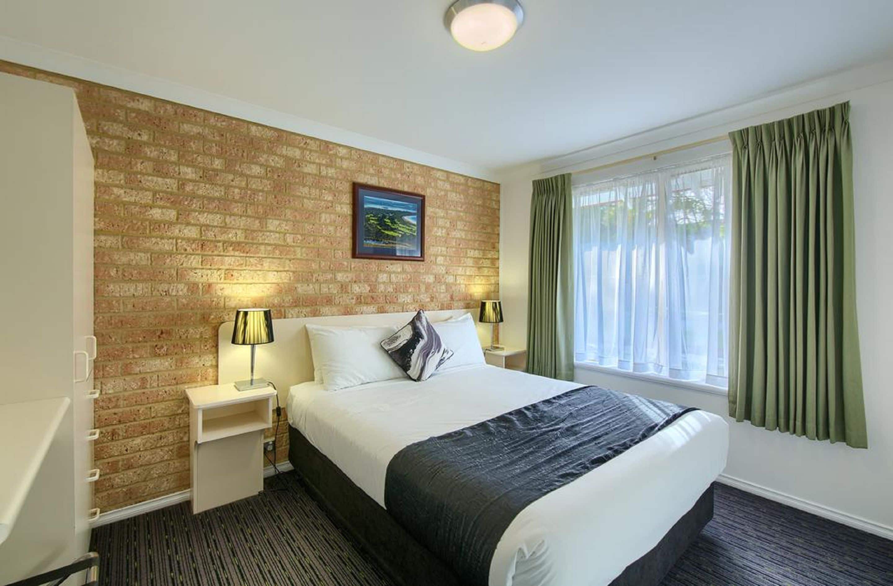 A hotel room with a brick accent wall, a large bed with a black and white throw, two bedside lamps, a nightstand, and a window with green curtains.