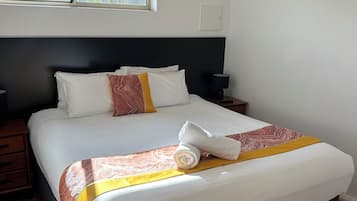 A neatly made bed with a rolled towel, a bedside table with a lamp, and a window with a view of trees.