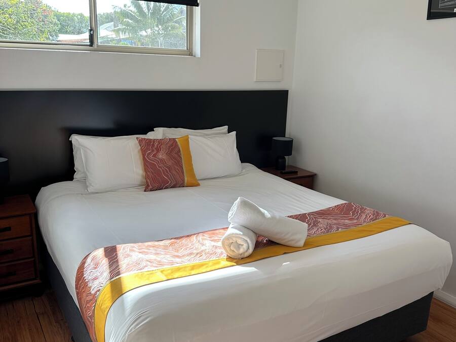 A neatly made bed with a rolled towel, a bedside table with a lamp, and a window with a view of trees.