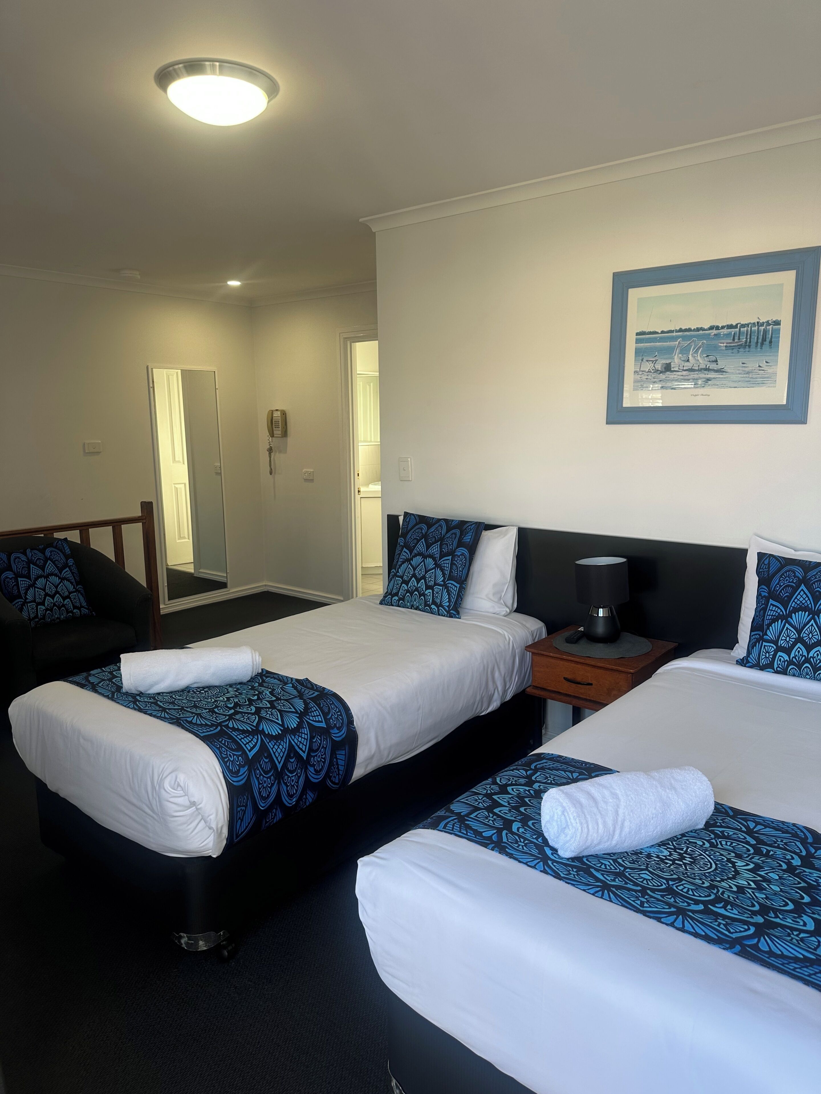 A hotel room with two beds, a bedside table with a lamp, a mirror, and a framed picture on the wall.
