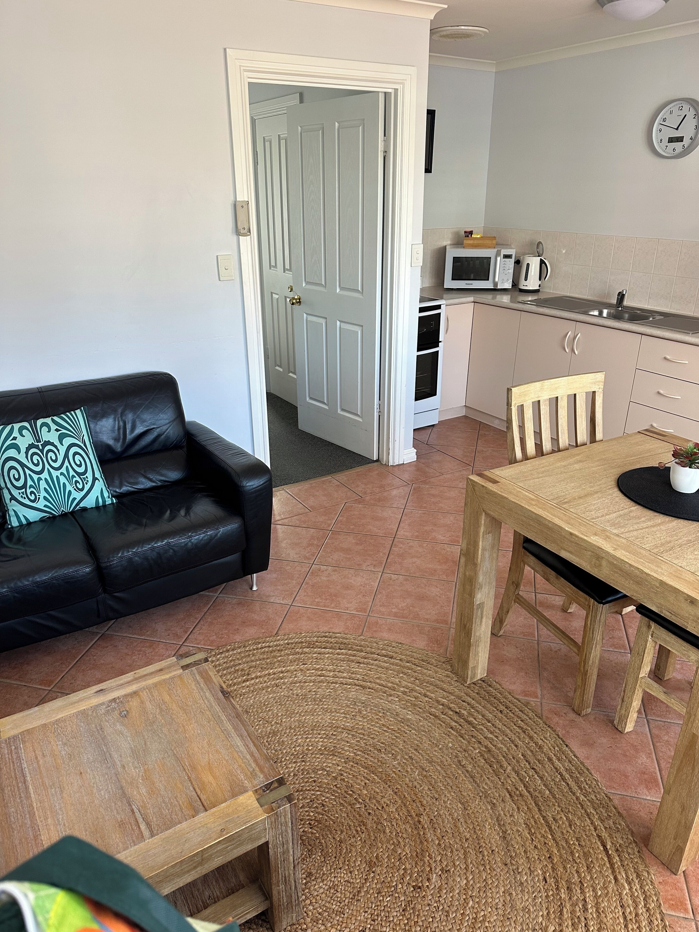 A living room with a black leather sofa, a wooden dining table, and a kitchen area with a microwave and oven.