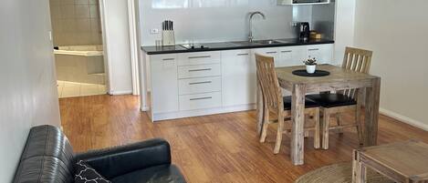A modern kitchen with white cabinets, a dining area with a wooden table and chairs, and a black leather sofa with a patterned pillow.