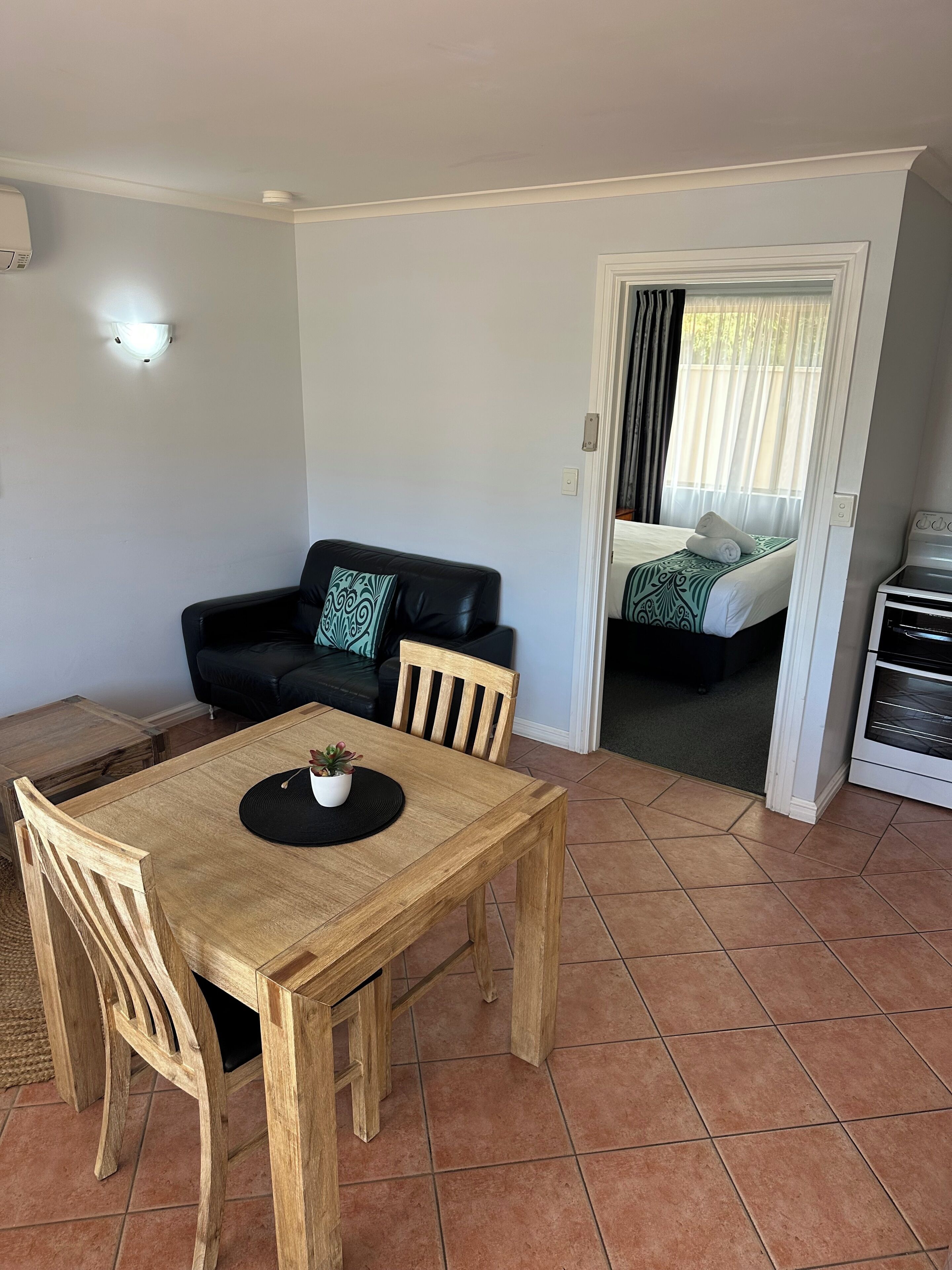 Uma área de refeições com mesa e cadeiras de madeira, um sofá de couro preto com almofada estampada, e uma cama com colcha verde e branca ao fundo.