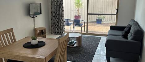 A living room with a wooden dining table, chairs, a sofa, a TV, and a sliding glass door leading to an outdoor area with a potted plant.