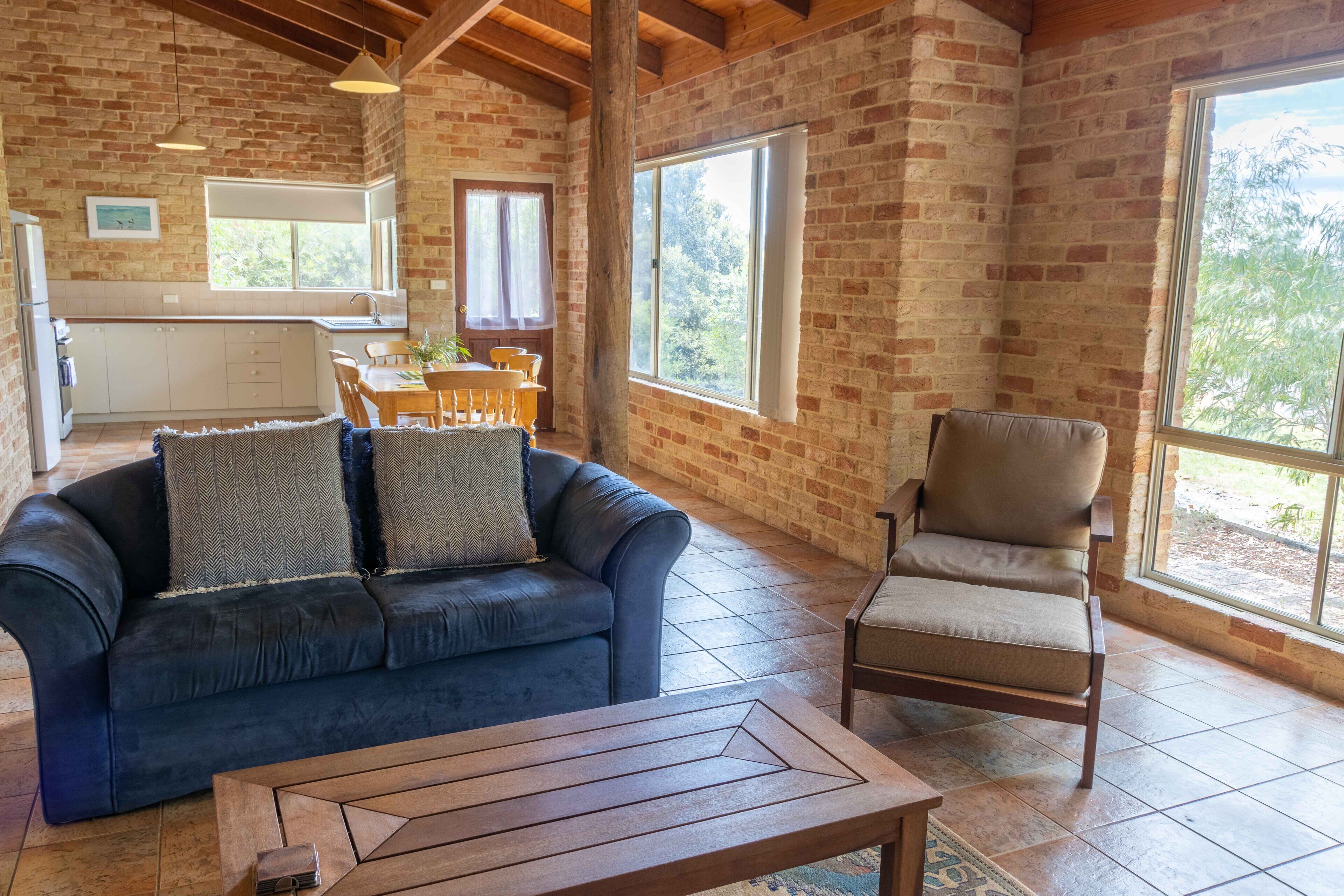 A spacious room with a brick wall, a wooden ceiling, a sofa, a chair, a dining table, and a kitchen area.