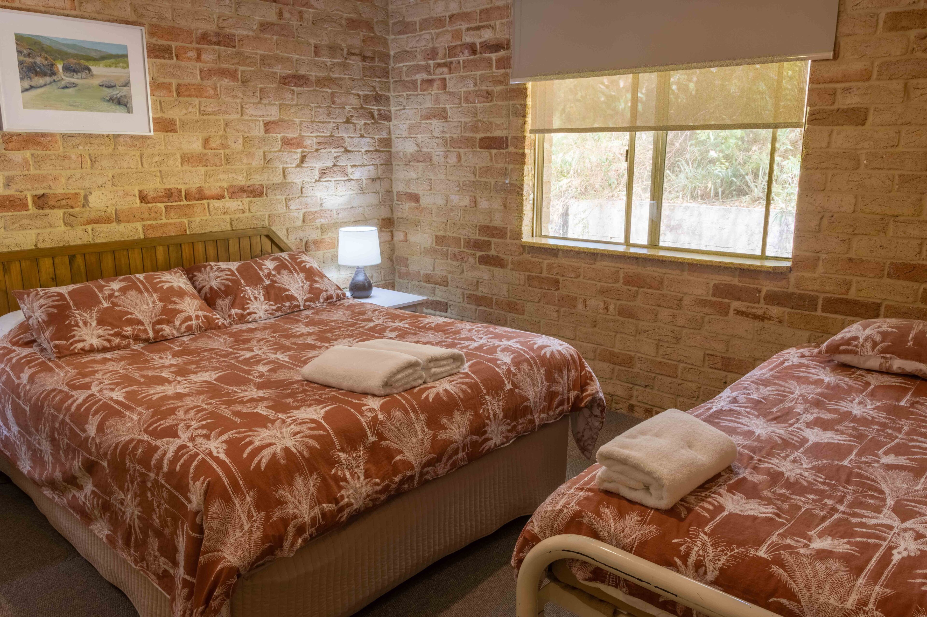 Ein Zimmer mit Backsteinwand, einem Bett mit blumigem Überwurf, einem Nachttisch mit Lampe und einem Fenster mit Blick auf Grünflächen.