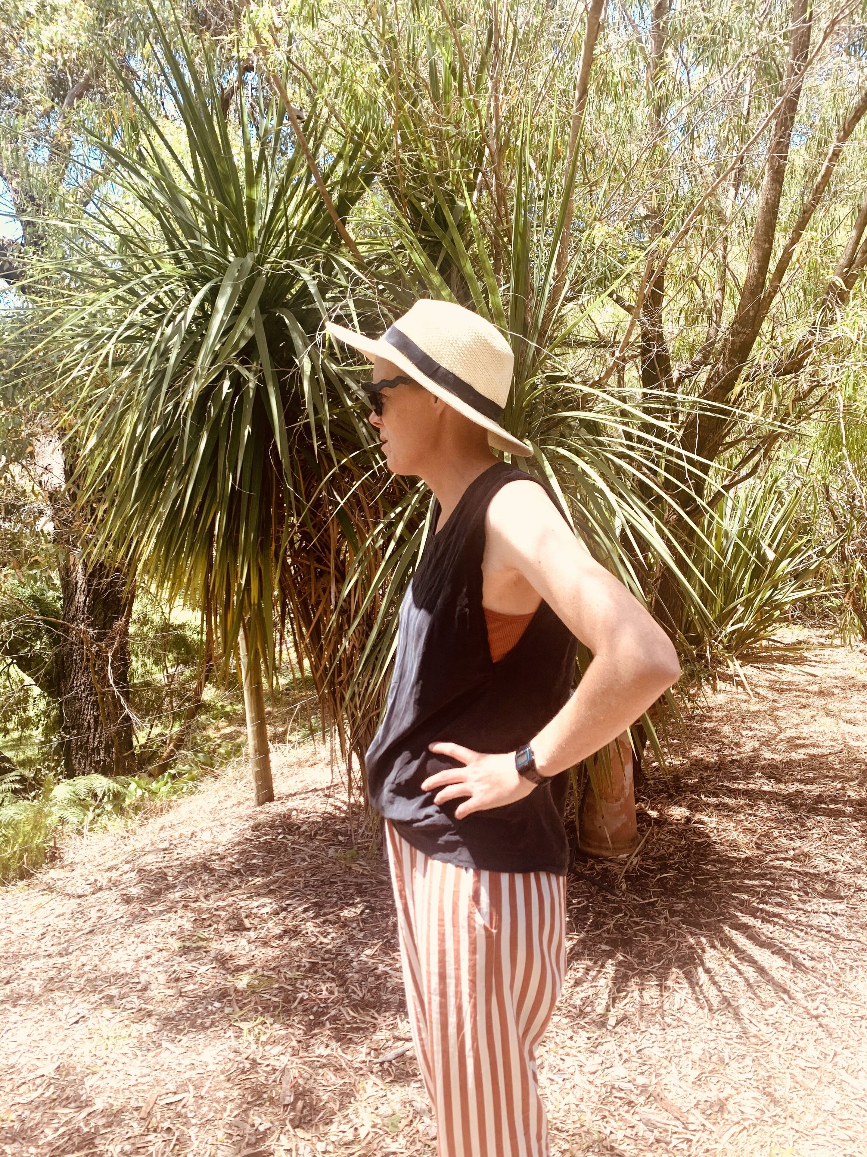Una persona parada al aire libre, con sombrero, vestida con una blusa negra y pantalones a rayas, rodeada de follaje.