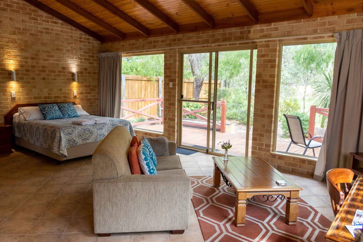 A bedroom with a bed, a sofa, a table, and a view of the outdoors through large windows.
