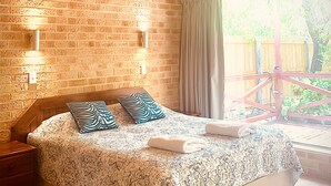 Ein Schlafzimmer mit Ziegelwand, Holzbalkendecke, einem großen Bett mit gemusterten Textilien, zwei Wandleuchten und Blick auf eine Holzterrasse und Grünfläche.