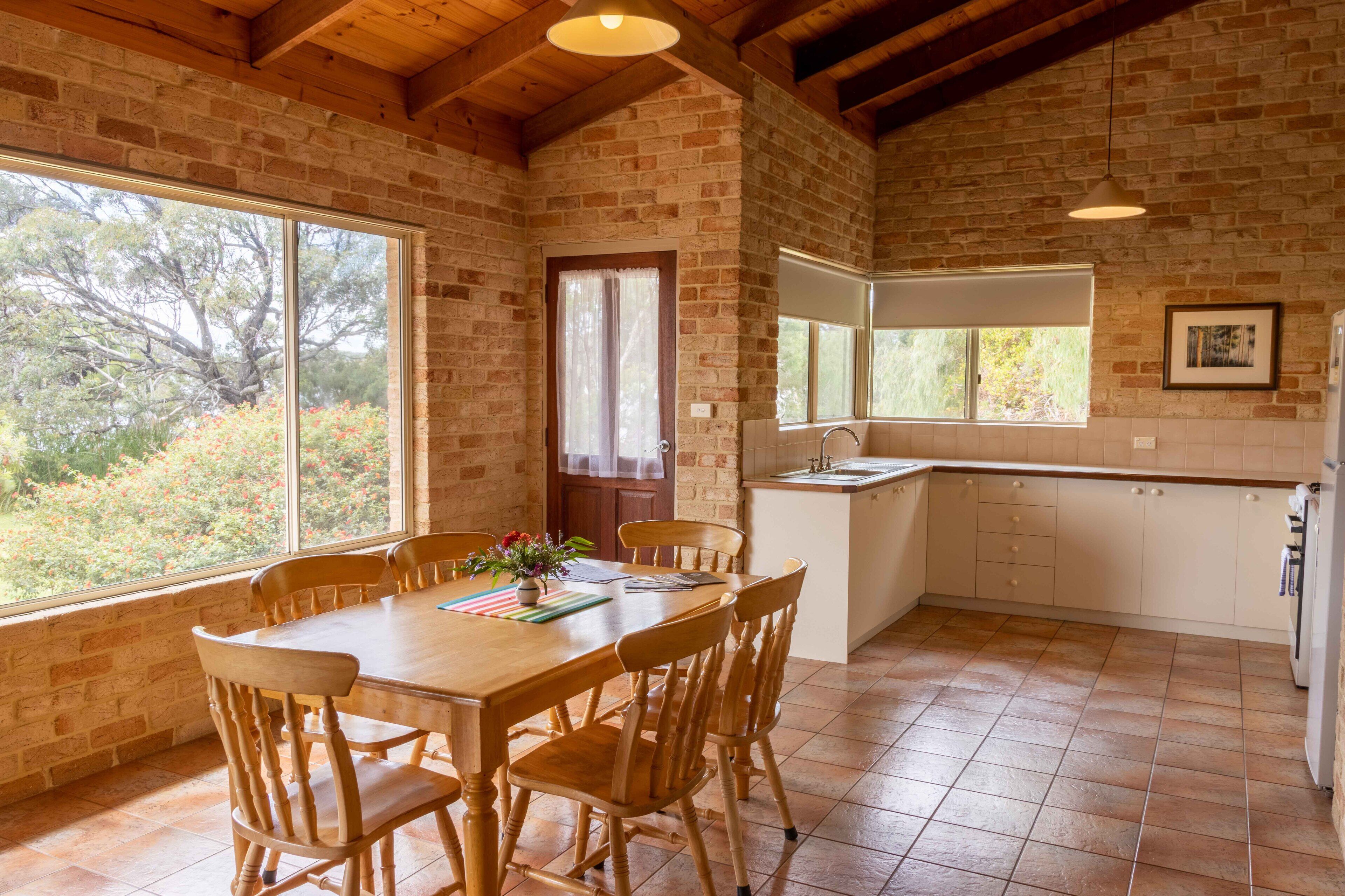 A rustic kitchen with a wooden table and chairs, a sink, and a window overlooking greenery.
