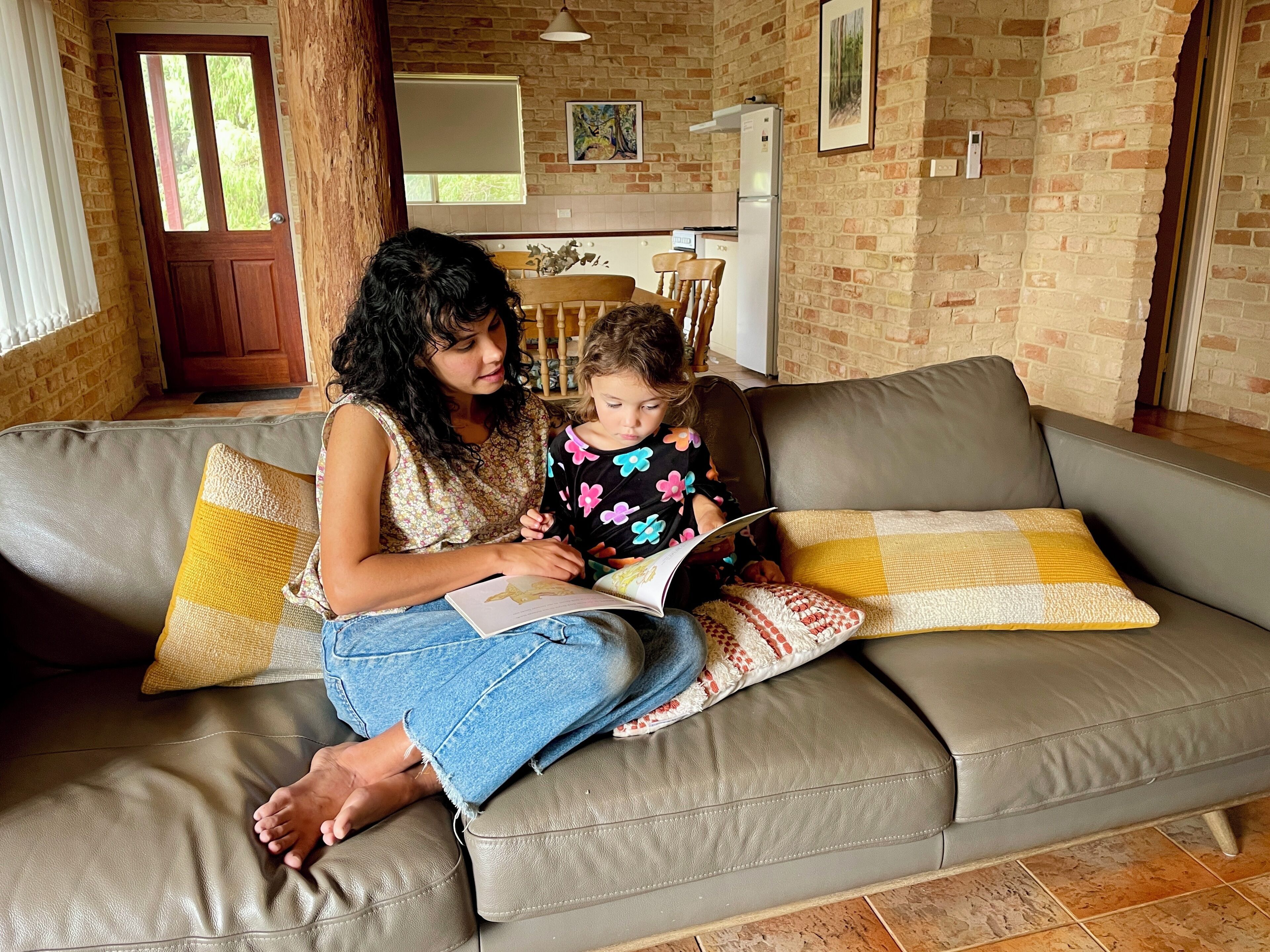 A woman and a child sitting on a leather couch with yellow and white cushions, reading a book.