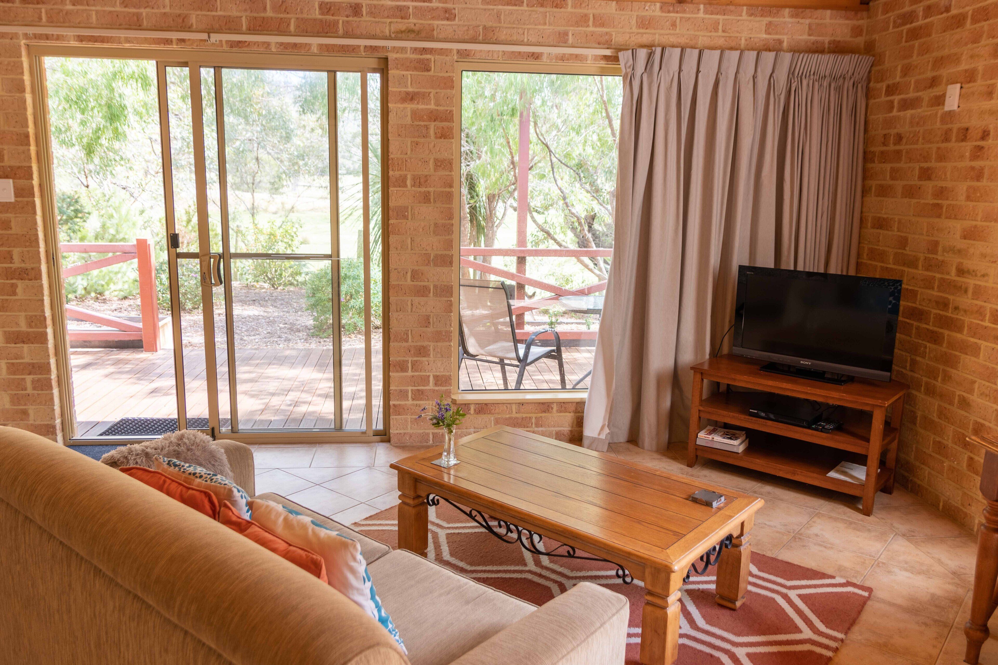 A living room with a sofa, a wooden coffee table, a TV on a wooden stand, and a sliding glass door leading to an outdoor patio with a table and chairs.