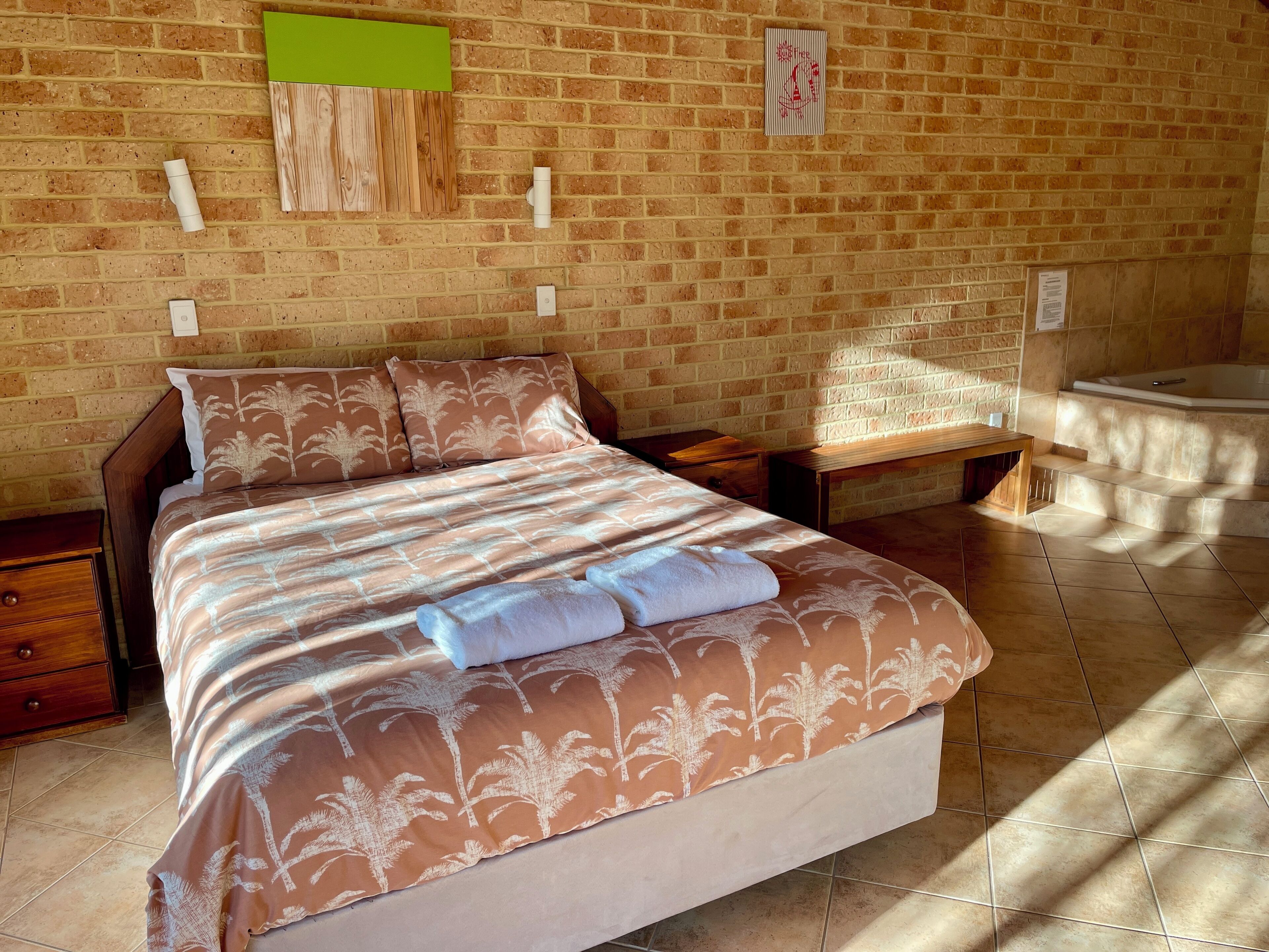 A neatly made bed with a patterned bedspread, a wooden nightstand, a bench, and a bathtub in a room with brick walls.