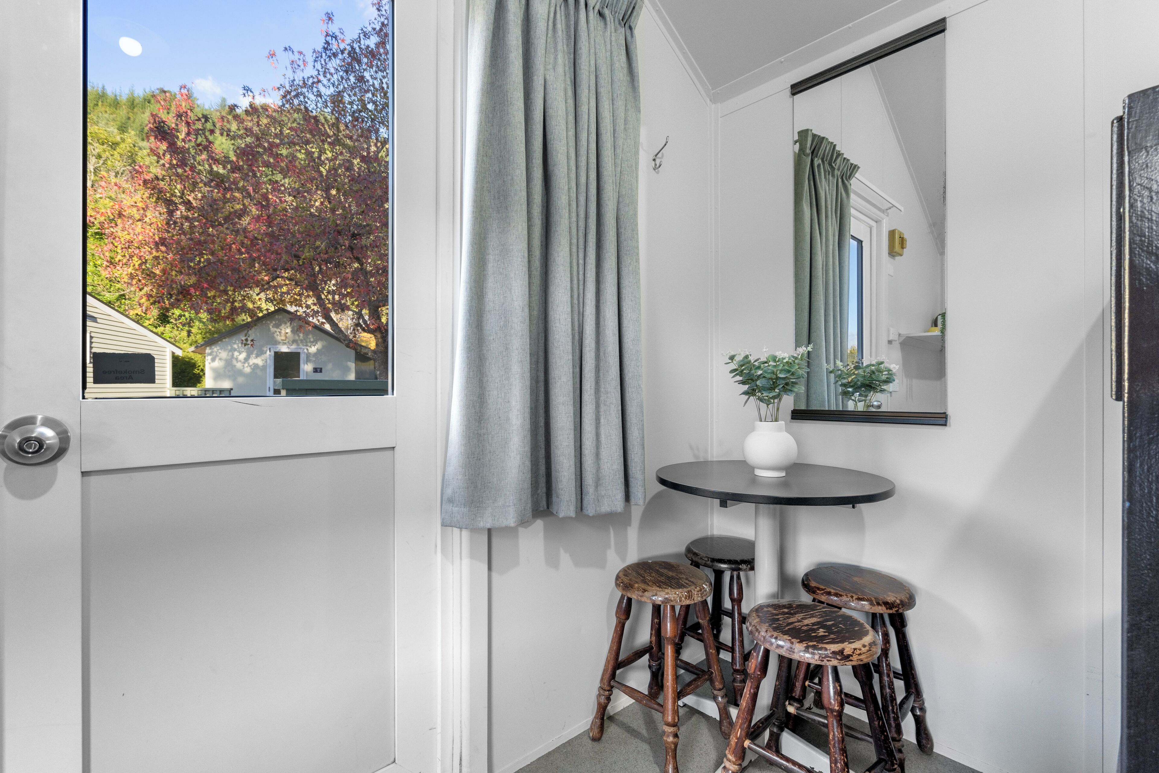 Una habitación con una puerta que da a un balcón, una mesa redonda con un florero y dos taburetes, y un espejo en la pared.