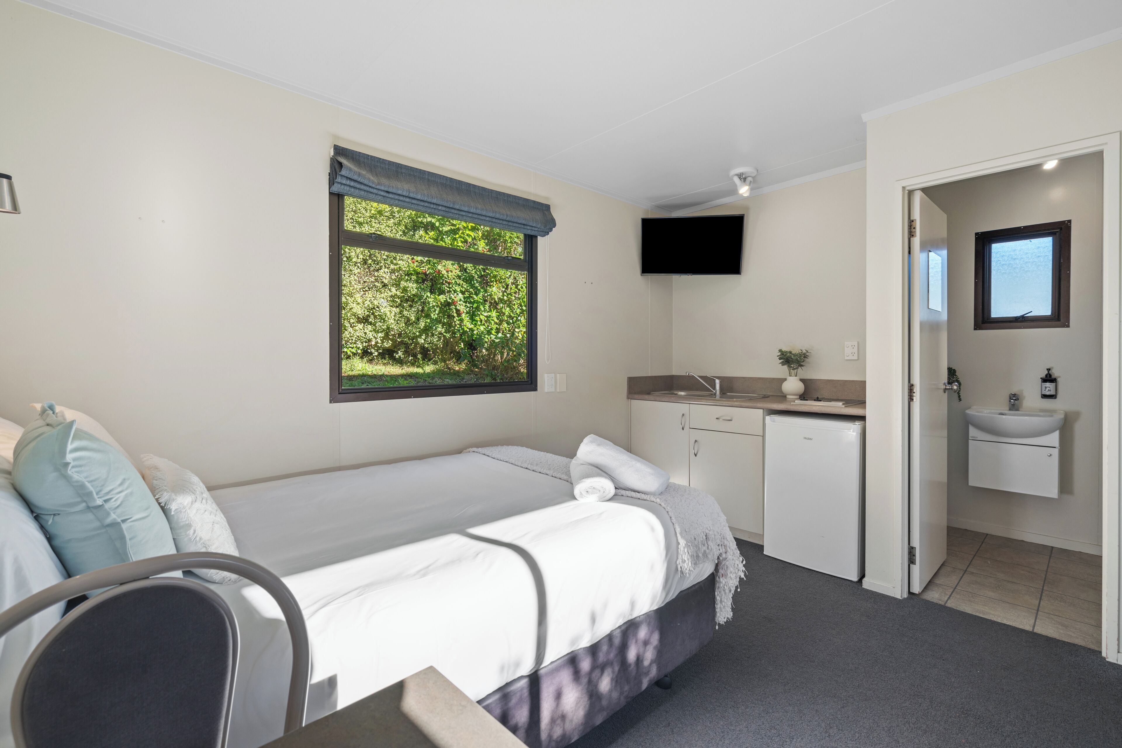 A bedroom with a bed, a desk, a window with a view of greenery, a TV, and a bathroom with a sink and mirror.