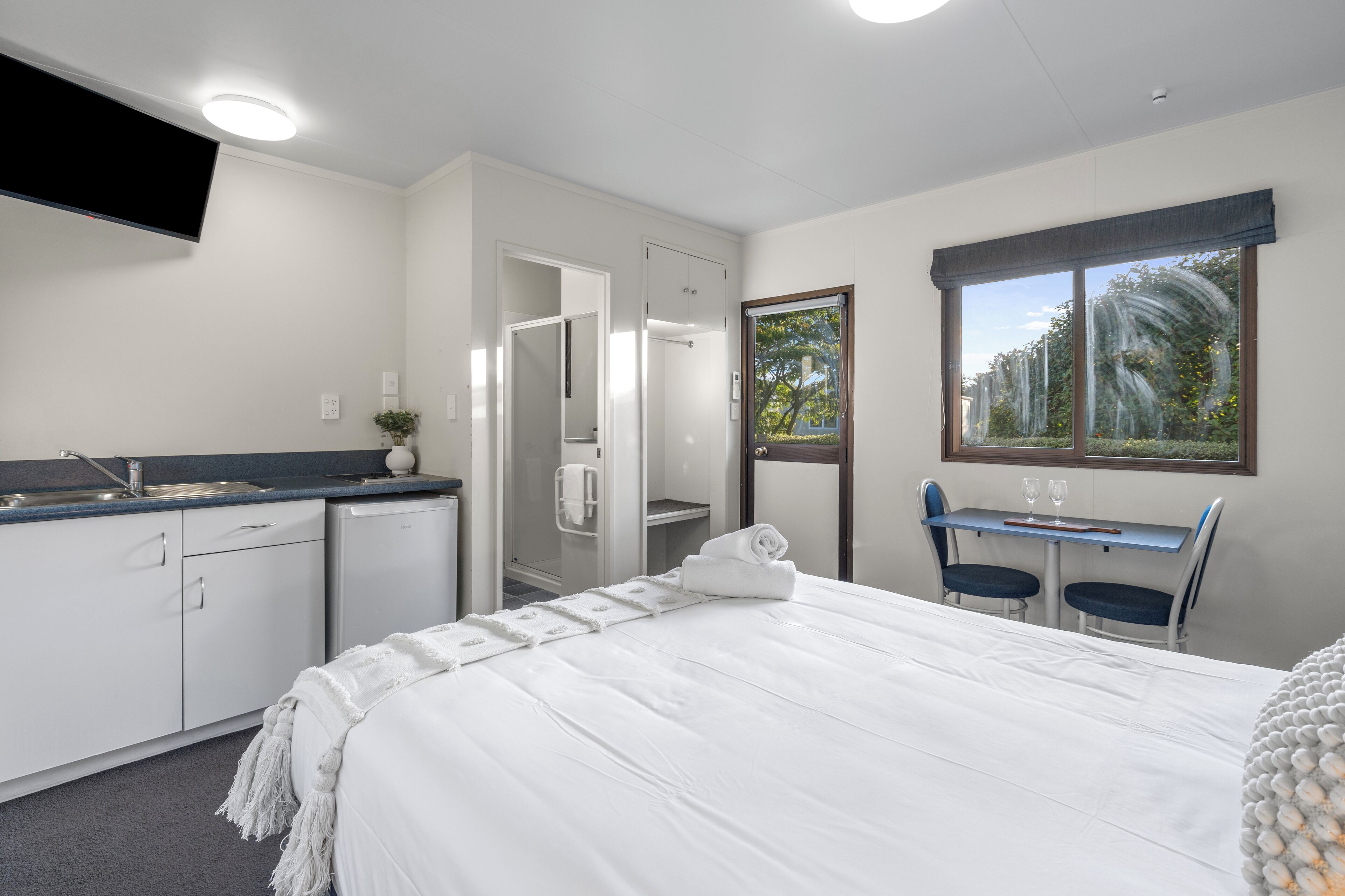 A hotel room with a bed, a desk, a chair, a TV, a shower, and a window.
