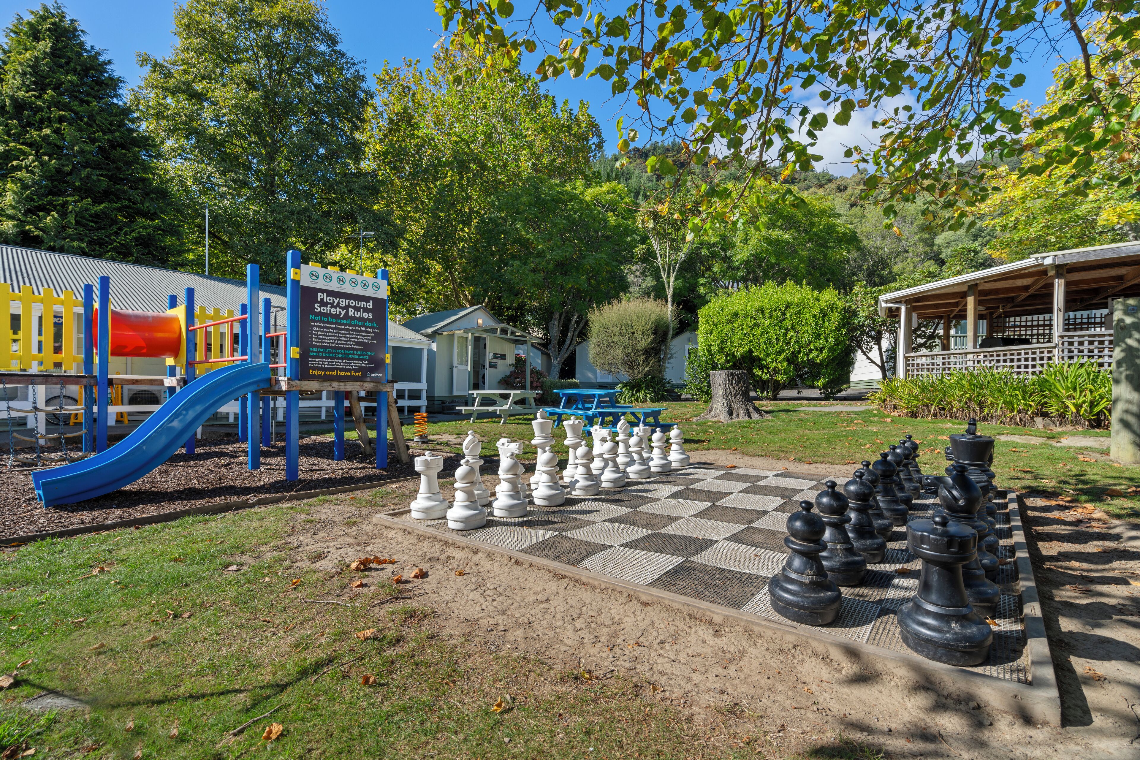 A playground with a slide and climbing structures, a chessboard with oversized chess pieces, and a picnic area with benches.