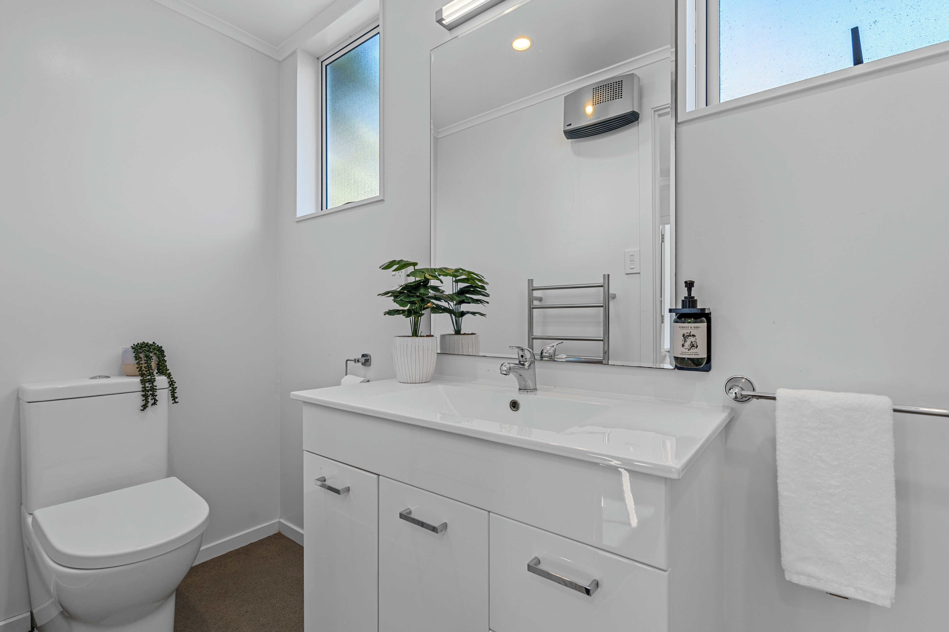 A modern bathroom with a white sink, mirror, and toilet. There are two potted plants on the sink counter.