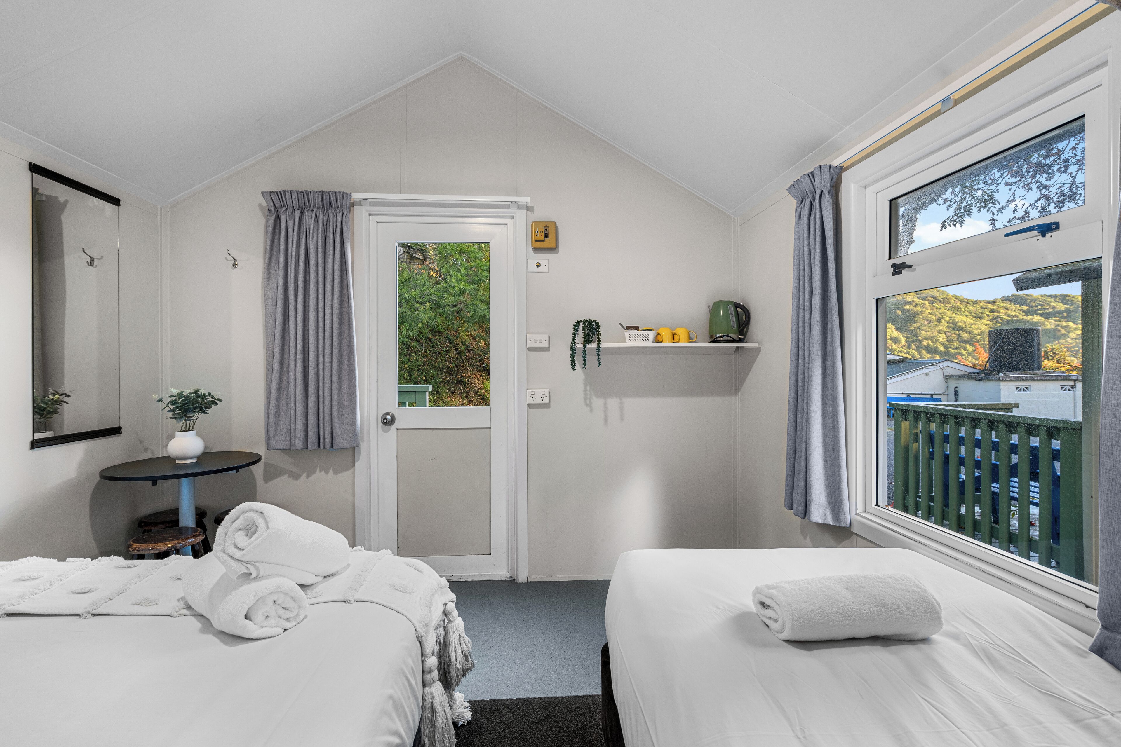 A bedroom with two beds, a small table with a plant, a window with a view of trees, and a door leading outside.