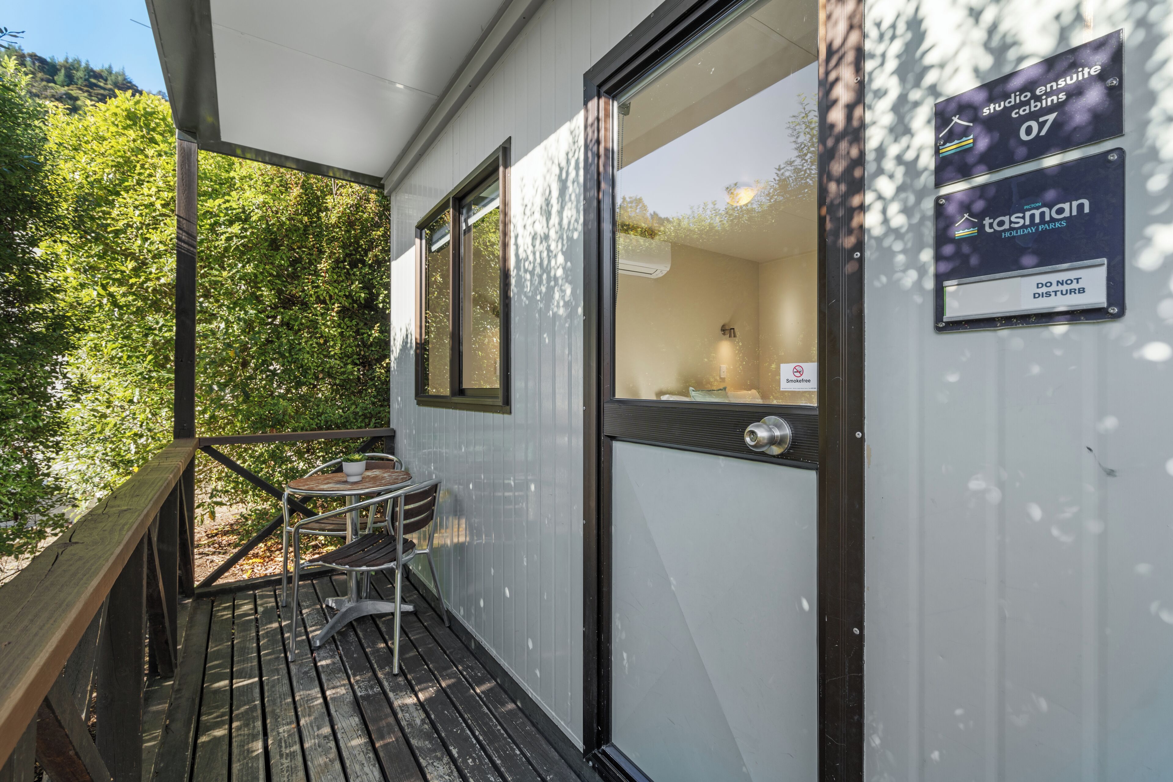 Terraza de madera con mesa y sillas, puerta de cristal y carteles que indican cabañas tipo estudio con baño privado y un cartel de 'No molestar'.