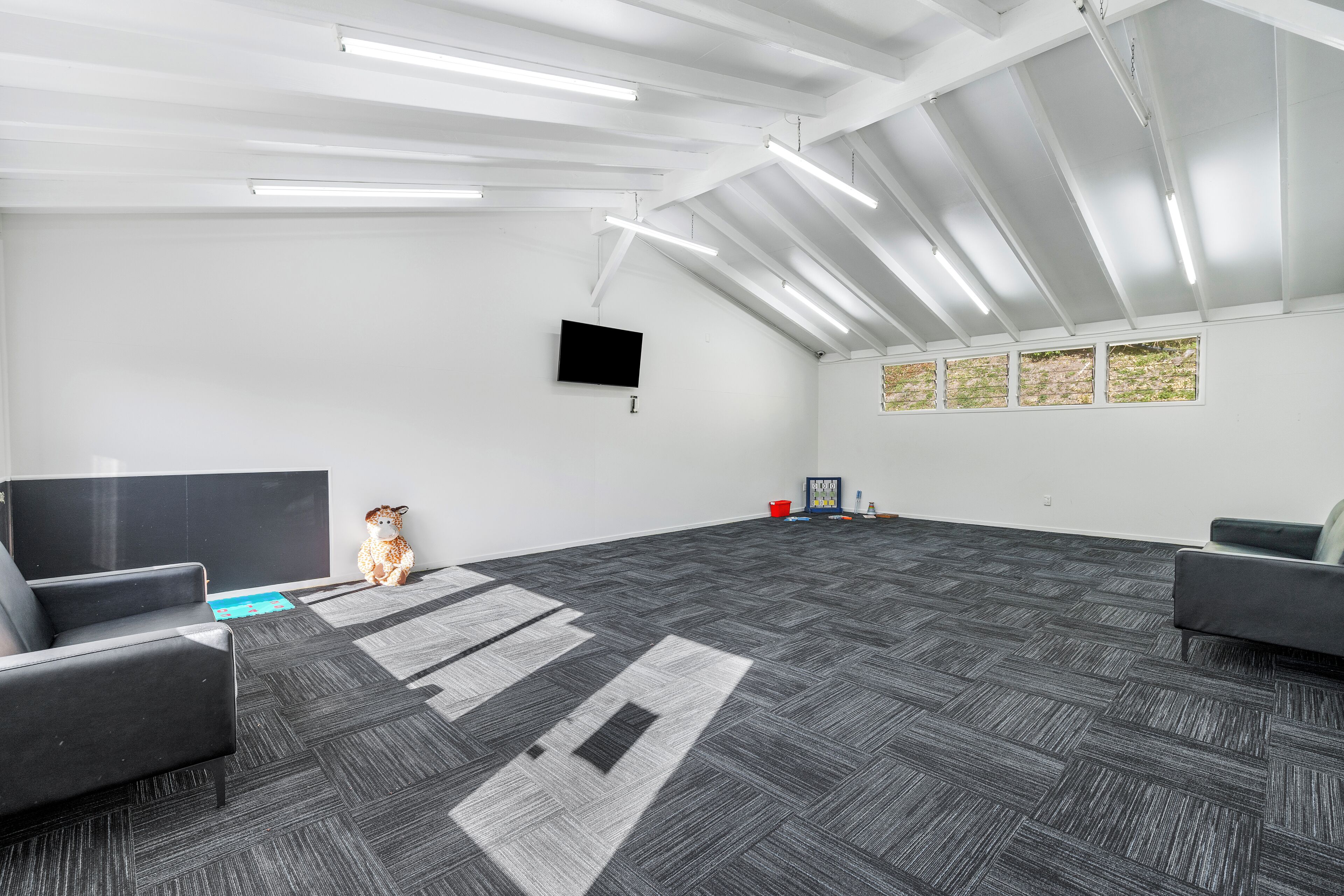 A spacious room with a large flat-screen TV mounted on the wall, grey carpet, and a ceiling with exposed beams and lighting fixtures.