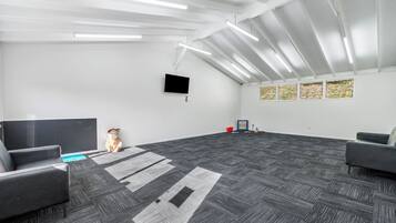 A spacious room with a large flat-screen TV mounted on the wall, grey carpet, and a ceiling with exposed beams and lighting fixtures.