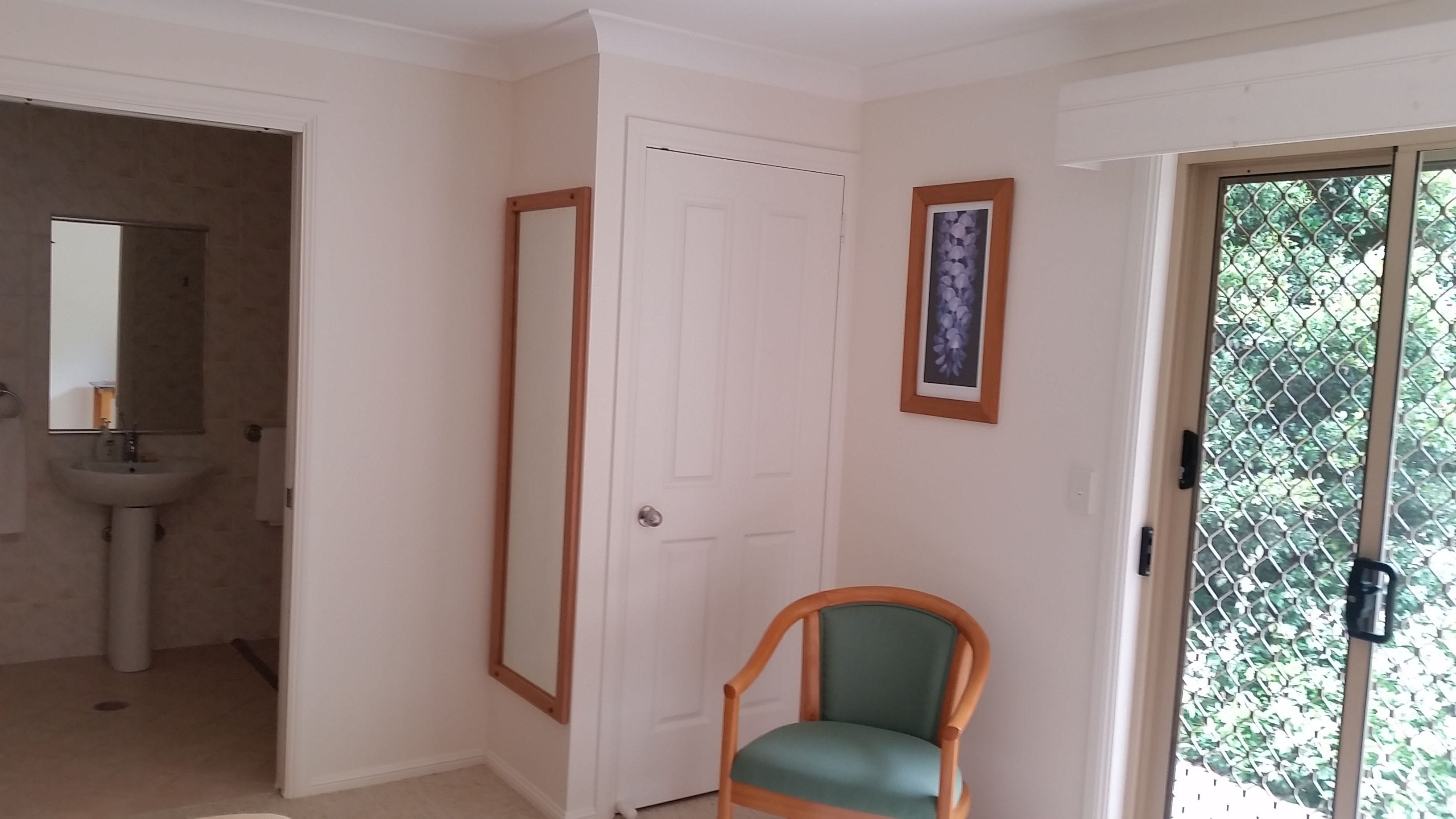 A room with a door, a framed mirror, a framed picture, a chair, and a window with a view of greenery.