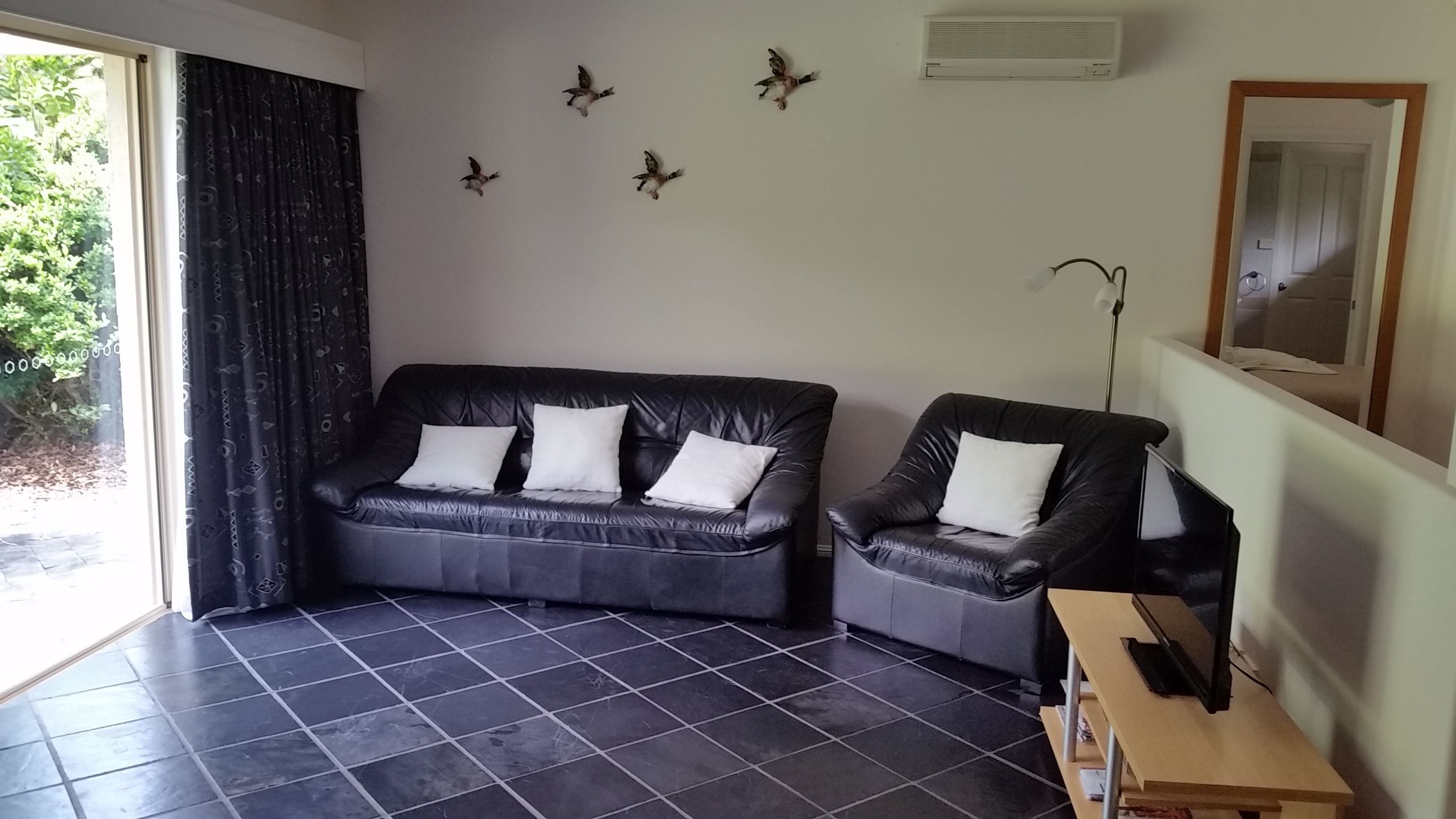 A living room with a black leather sofa and armchair, a wooden side table, and a large window with curtains.