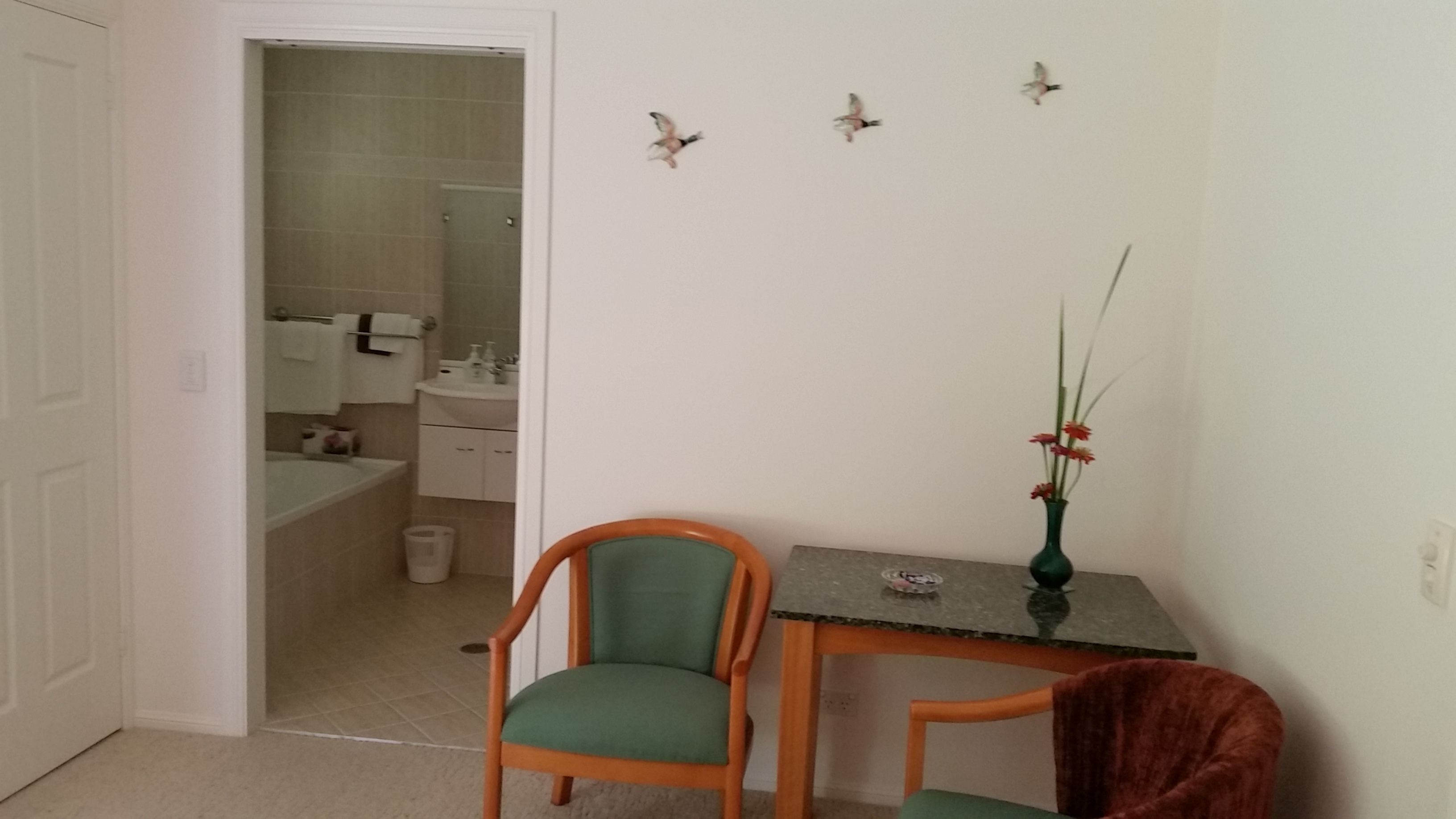 A small room with a green chair, a wooden table, and a vase with flowers. There is a door leading to a bathroom.