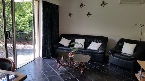 A modern living room with a black leather sofa, a glass coffee table, and a view of greenery outside. - Mescal's at Pampoolah B&B (Pampoolah)
