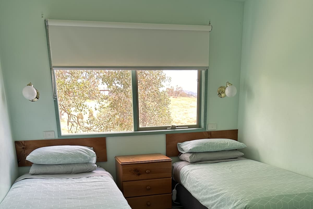 A room with two beds, a wooden nightstand, a window with a view of trees, and a ceiling fan.
