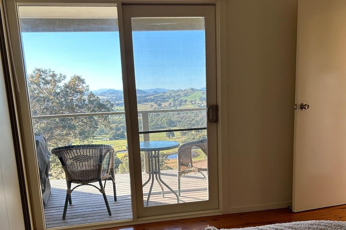 A bedroom with a large bed, a view of a balcony with outdoor furniture, and a door to the right.