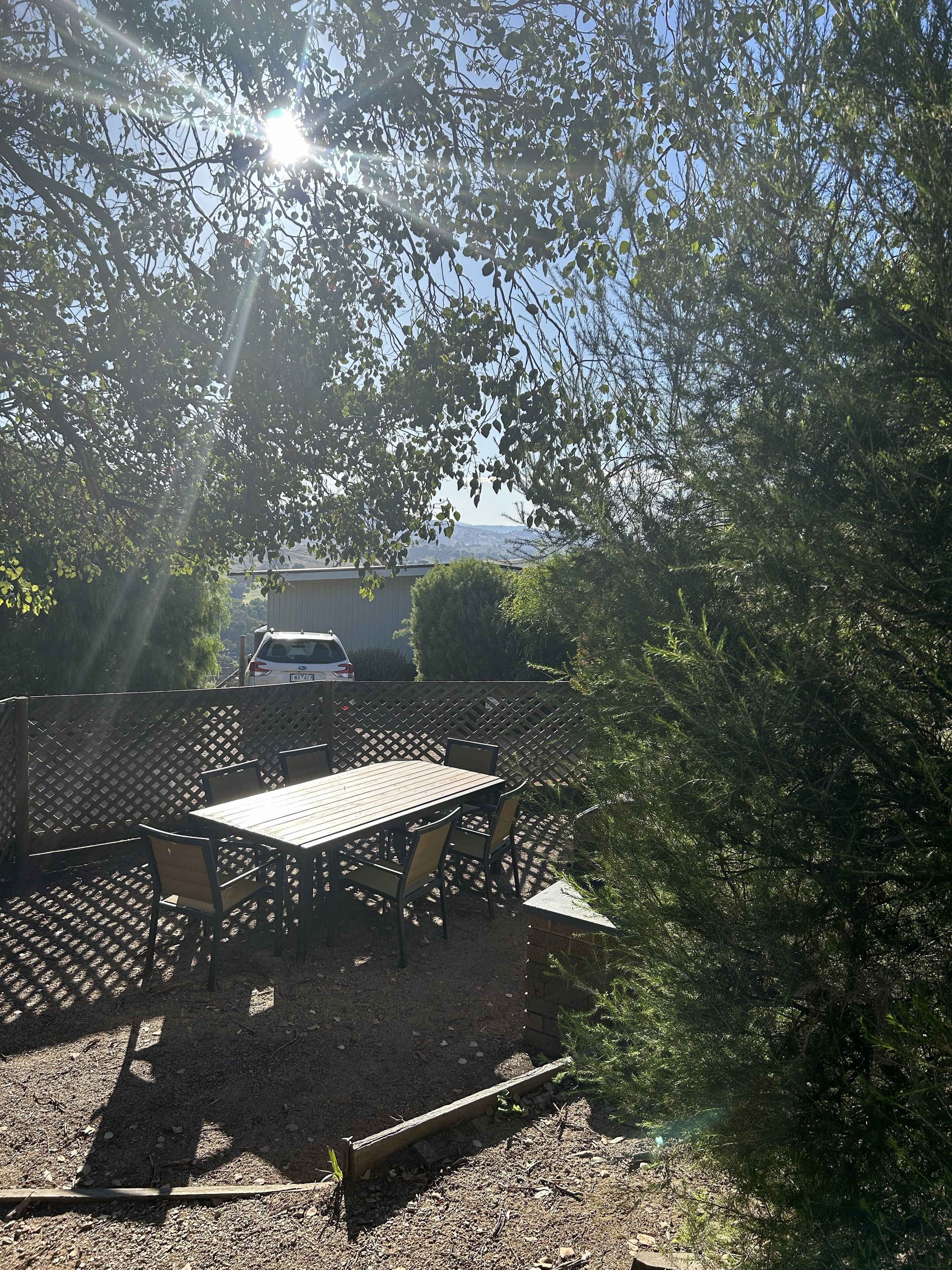 A patio area with a wooden table and chairs, surrounded by a lattice fence, and a car parked in the background.