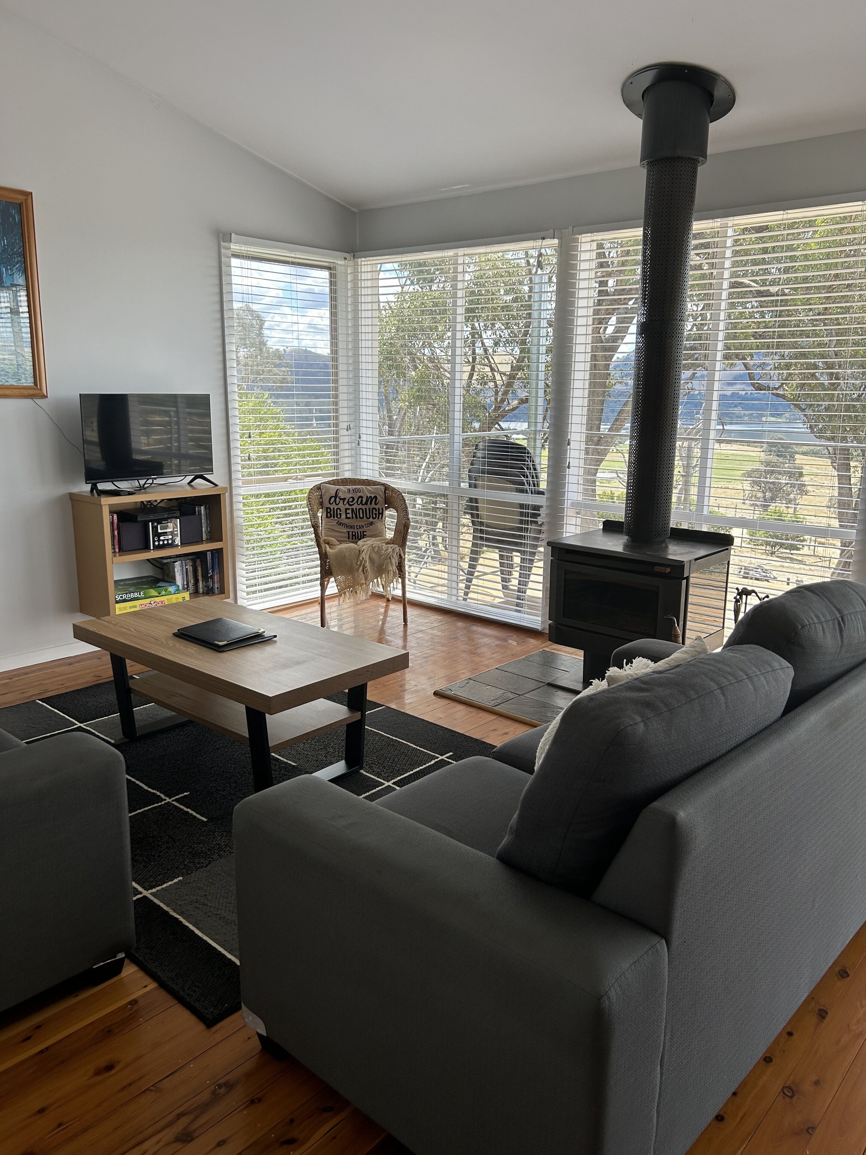 A modern living room with a sofa, a coffee table, a TV on a stand, a bookshelf, and a large window with blinds.
